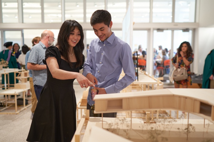 Two smiling students discuss a wooden model of a long, low building on stilts.