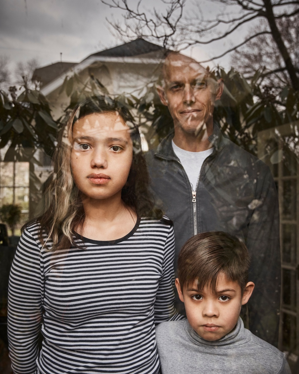 A photograph of two tanned children, one female and one male, with an older man standing behind them looking out a window with a reflection of a house and tree on it.