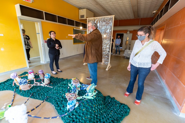 Professor Jamie Adams speaks to Karen Yung in front of her soft sculpture installation on the ground. The sculpture has 8 figures that sit on a dark green mount connected to another two small figures on the left