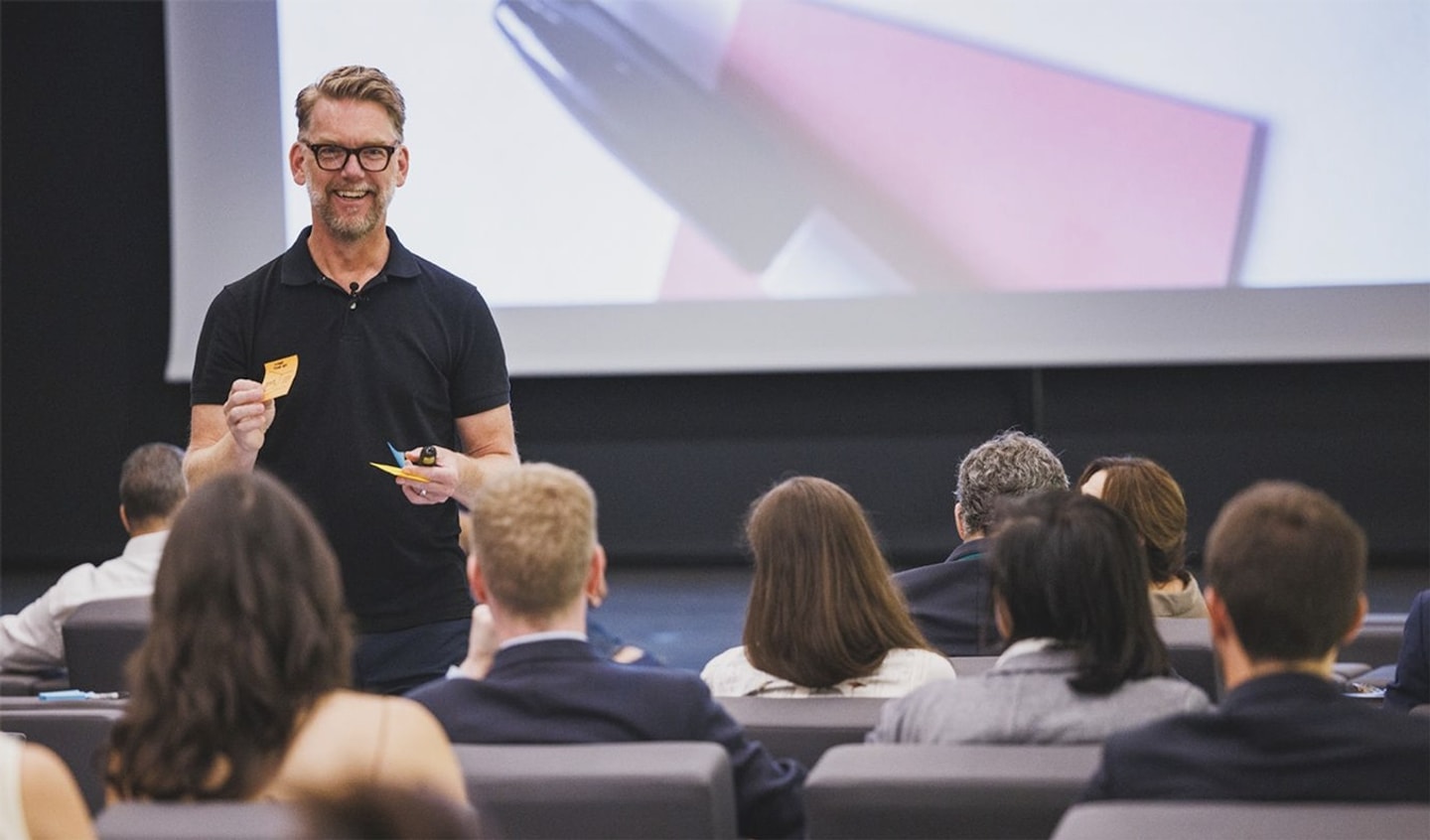 A man in a black polo shirt and glasses holds sticky notes while talking to a seated audience.