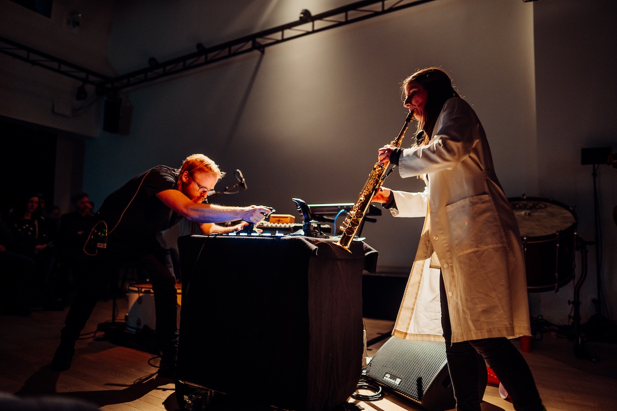 Two people perform on a spotlit stage. One on the left bends over a table manipulating a machine while on the left a woman in a long white coat plays a long brass wind instrument. 
