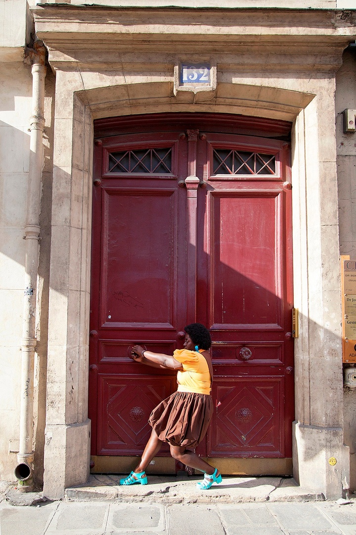 Photograph of an individual in a gold shirt, brown skirt, and teal shoes pulling on one knob to a huge, burgundy double door that is framed by stonework.