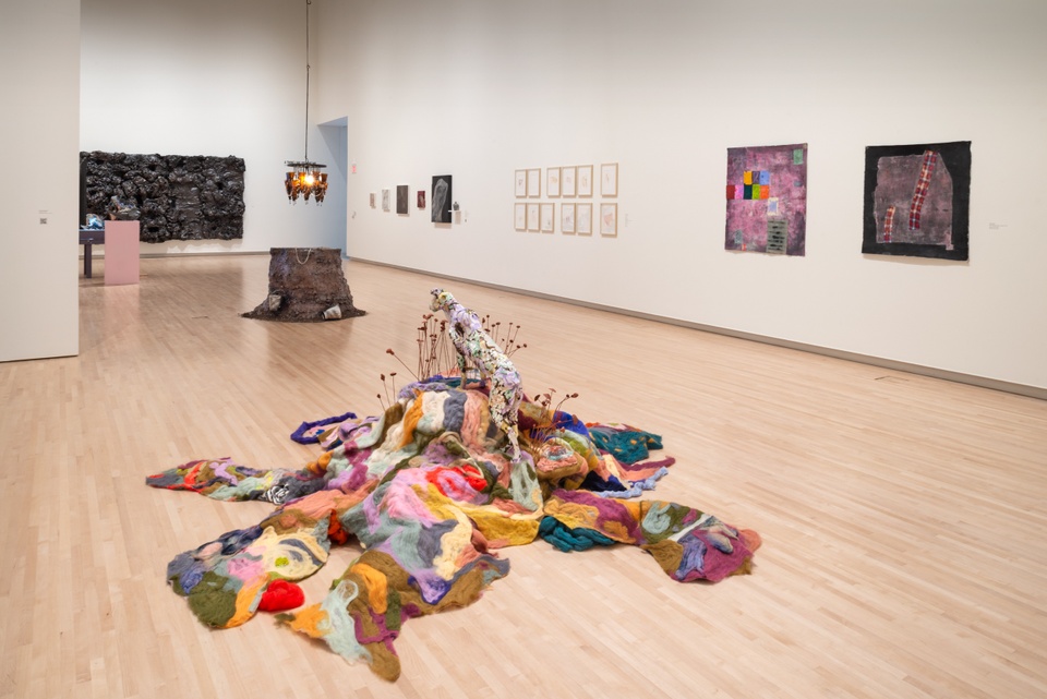 Overview of gallery showing a soft colorful felt sculpture on the floor, a tree stump with a glass chandelier, and two paintings on the wall.