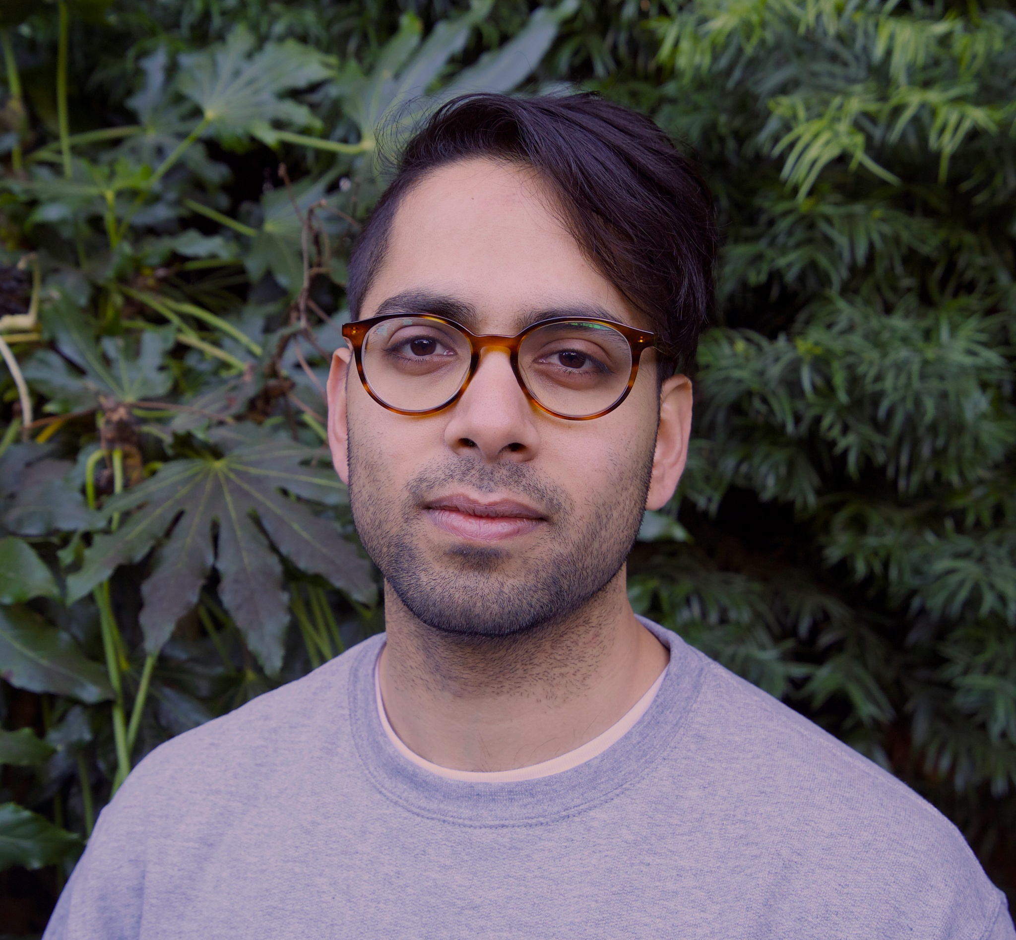 A headshot of Yusuf Siddiquee posing against a background of dark green leaves. He wears a lilac crewneck sweater and glasses. He looks gently at us. 