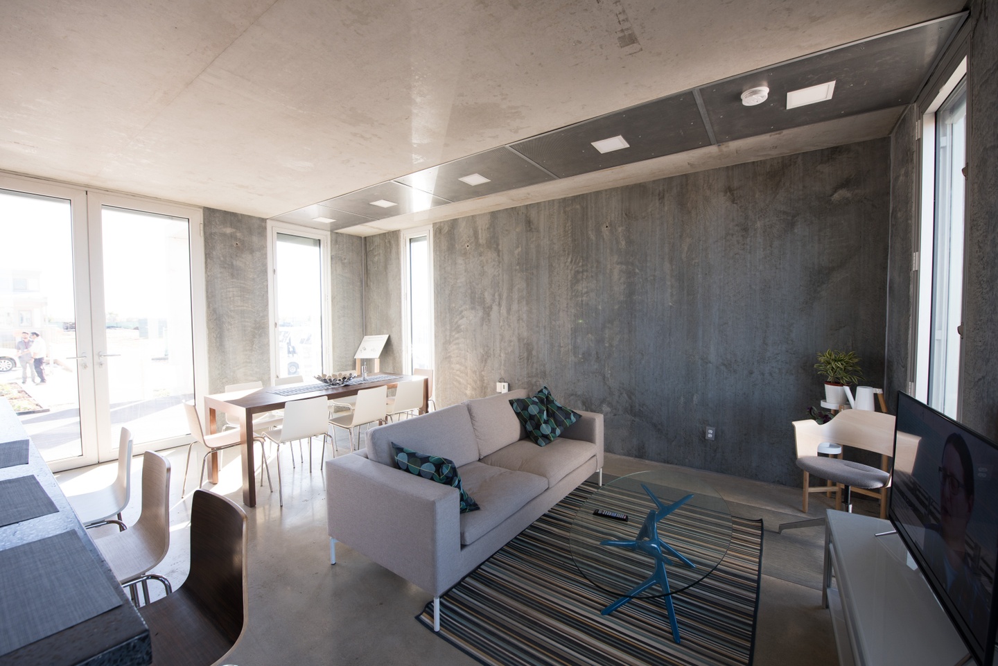 Interior of a house with concrete floors, walls, and ceiling with several full-height windows and furnished with neutral-toned modern furnishings.