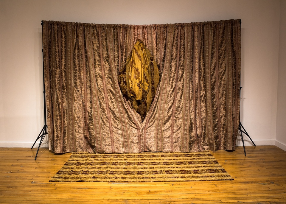 A curtain made of brown shiny upholstery fabric with a yellow, bunched segment the shape of a vulva in the center.