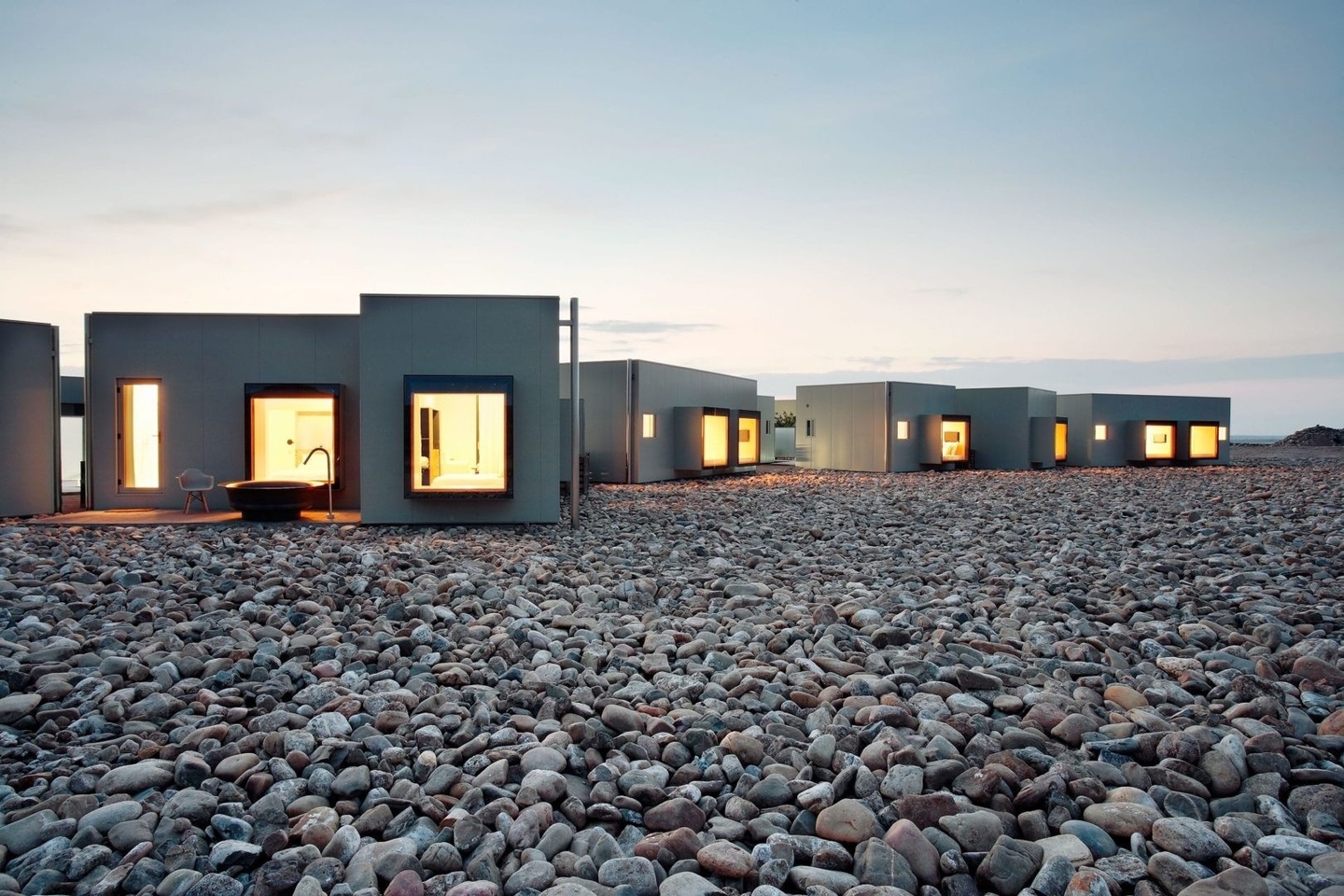 Photo of a contemporary, single-story hotel with gray, smooth walls interspersed with bumped out, square-shaped windows, the closest to the righthand of the frame revealing warm light and a bed. The hotel is built atop a bed of rocks.