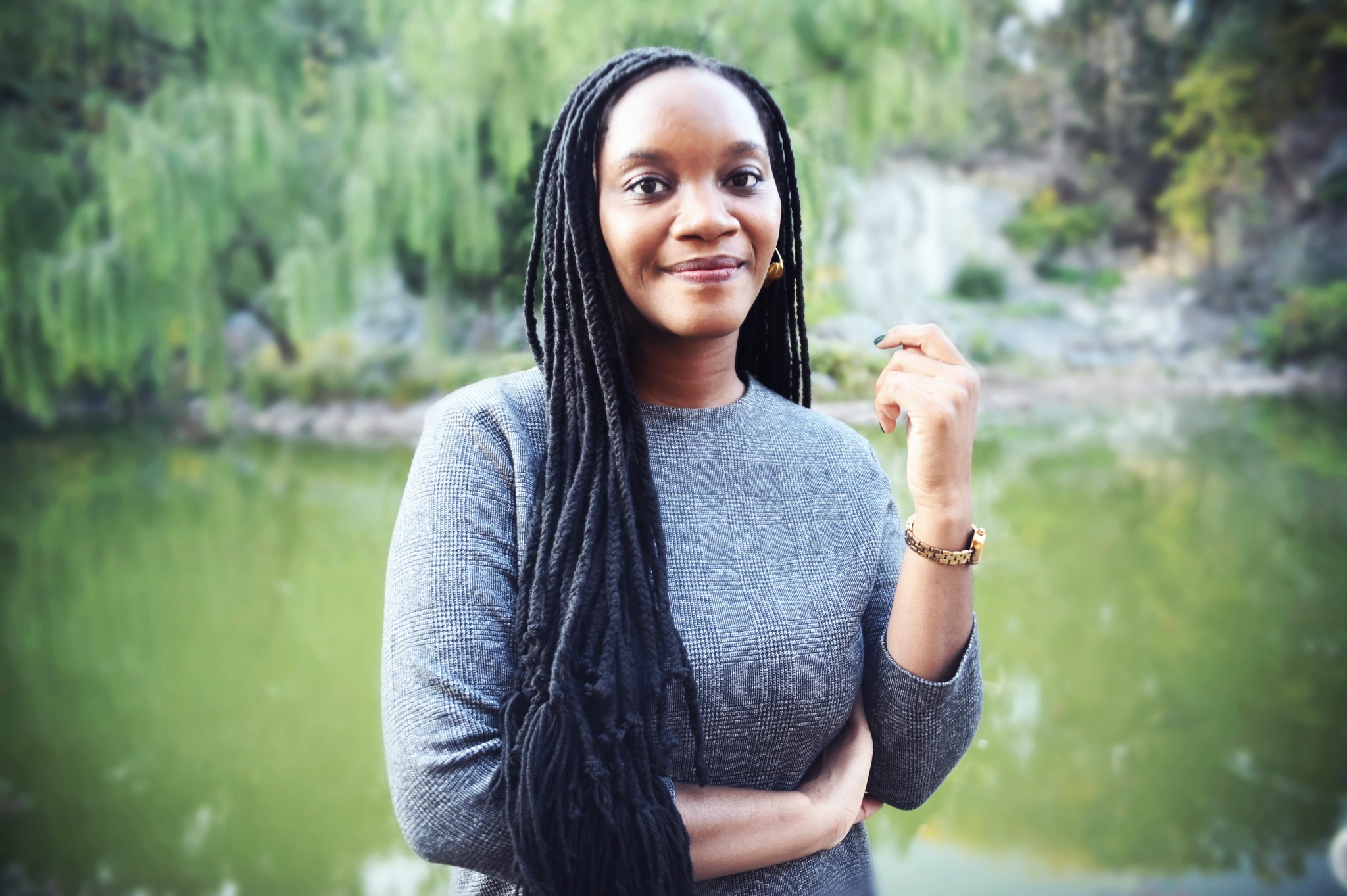 A photo of the artist Ladi'Sasha Jones who poses with a pond in the background. Jones has one hand raised and one crossed across her waist, with long hair falling over her right shoulder. 
