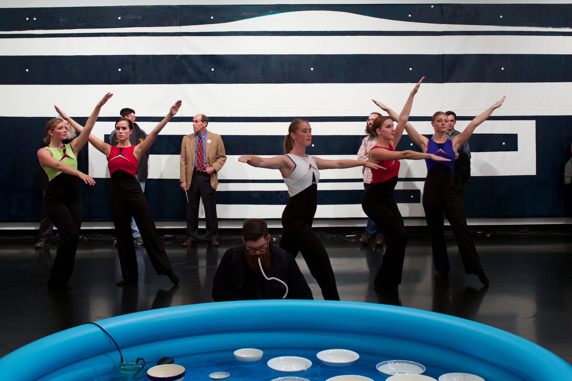 A group of light-skinned, female dancers pose in front of a black and white painted wall with a group of people looking on. In front of the dancers, there is a blue, blow-up pool with white bowls floating in it and a man crouching by it.