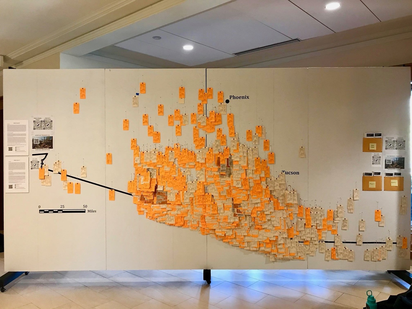 Installation of thousands of orange and white toe tags on white display boards, with black labels for cities like Phoenix and Tucson as well as a mileage key.