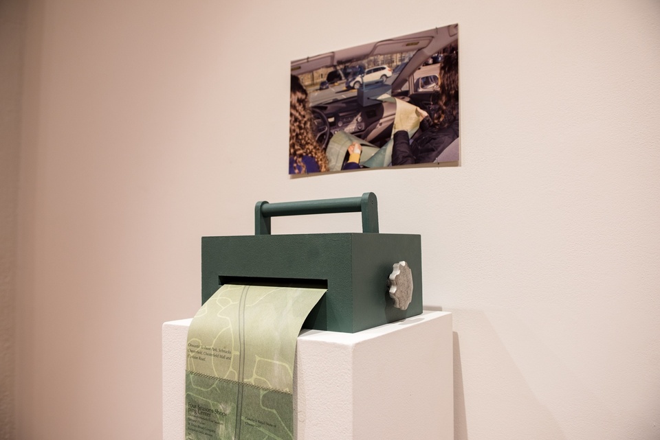 A green wooden box with a handle the size of a desktop printer sits on a plinth. A strip of green hand-made paper spits out of a slot in the side. Above it on the wall is hung a photo of two people in a car with this box, inspecting the very long sheet of green paper.