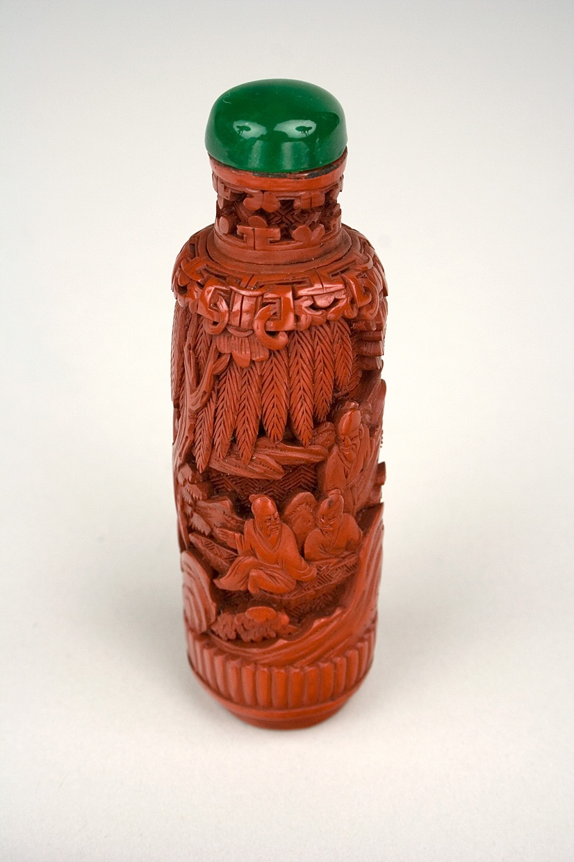 A small, cylindrical, orange bottle carved with images of plants and men sitting cross-legged with a green top.