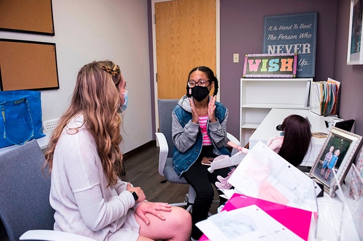 Two seated people wearing Covid masks are in conversation, one with hands held up about six inches apart.