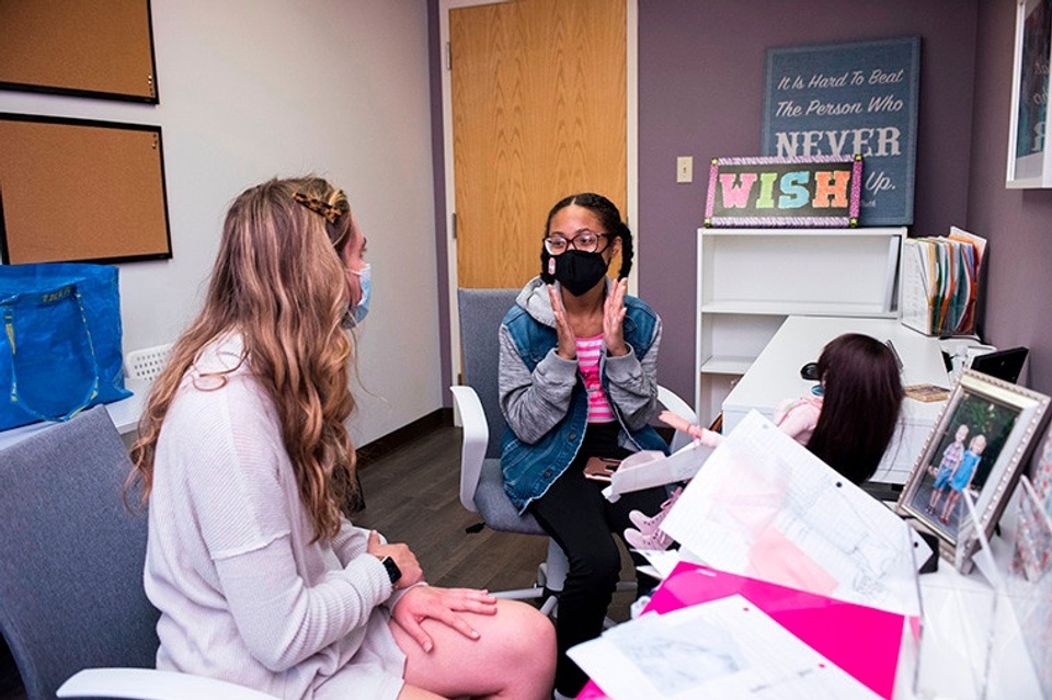 Two seated people wearing Covid masks are in conversation, one with hands held up about six inches apart.