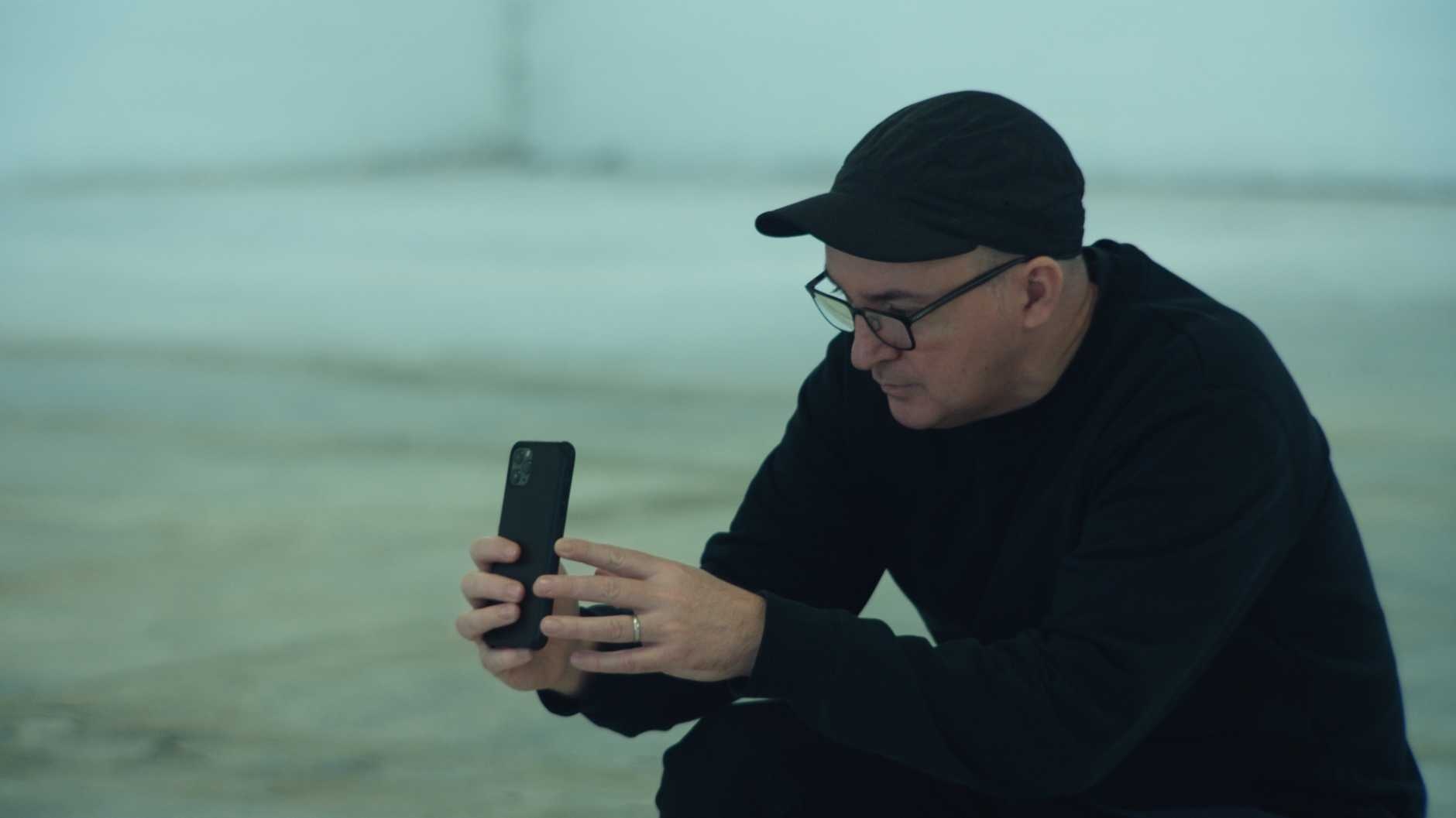 A white man in a dark gray baseball cap and glasses kneels to steady an iPhone that he concentrates on as if taking a photo with it.