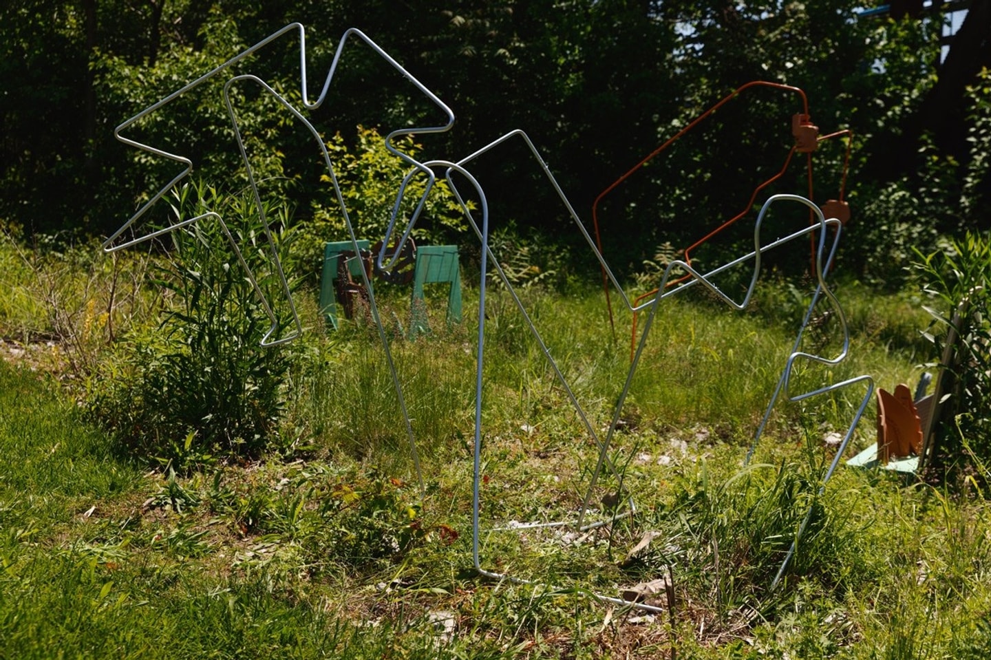 An outdoor sculpture installation by Ribaudo featuring silver and orange bent-wire geometric outlines standing in a sunny, grassy field framed by dense trees