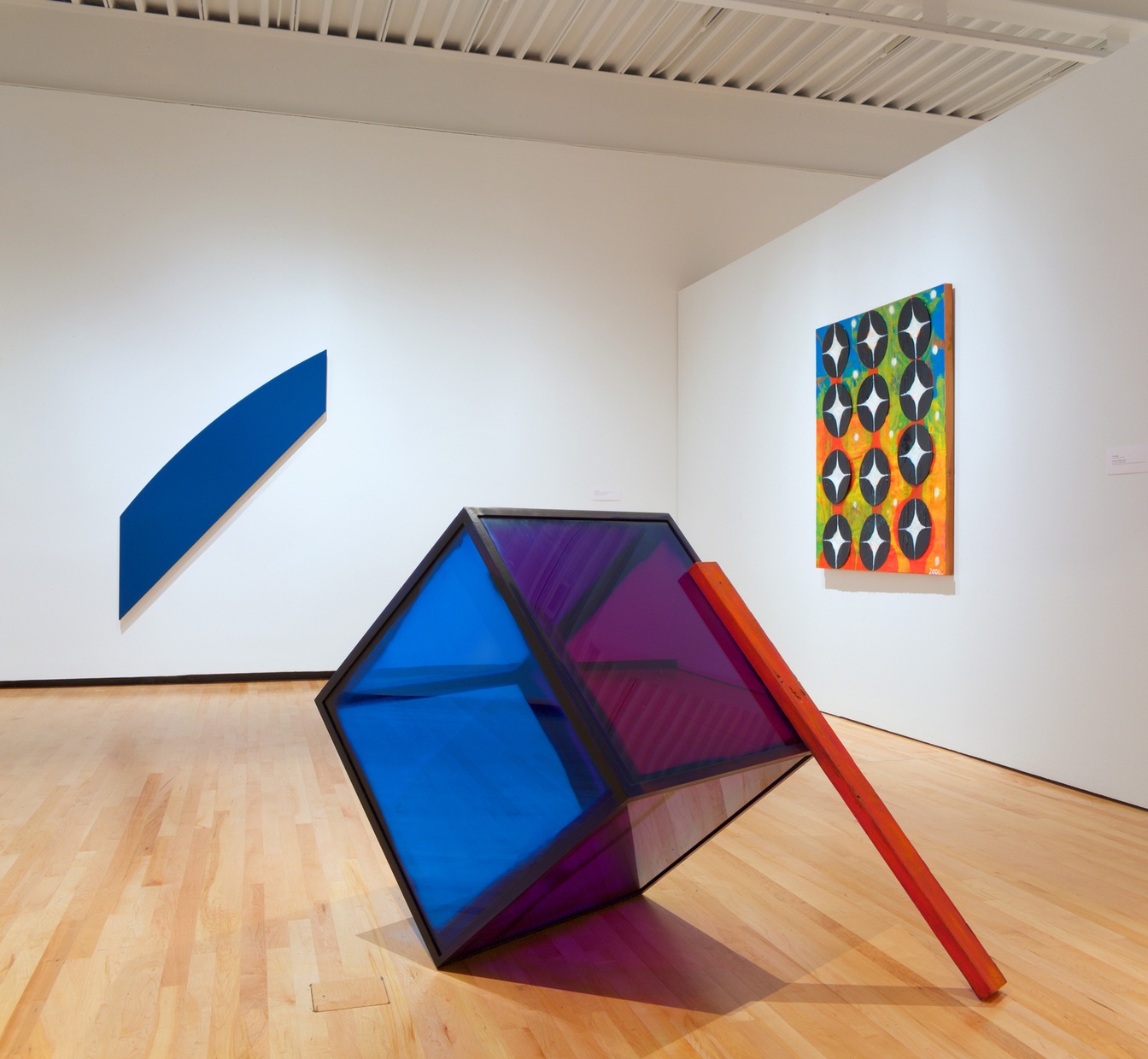 A large, purple and blue, translucent cube that is tilted at an angle with a red stick leaning against it and two abstract artworks on the background walls.
