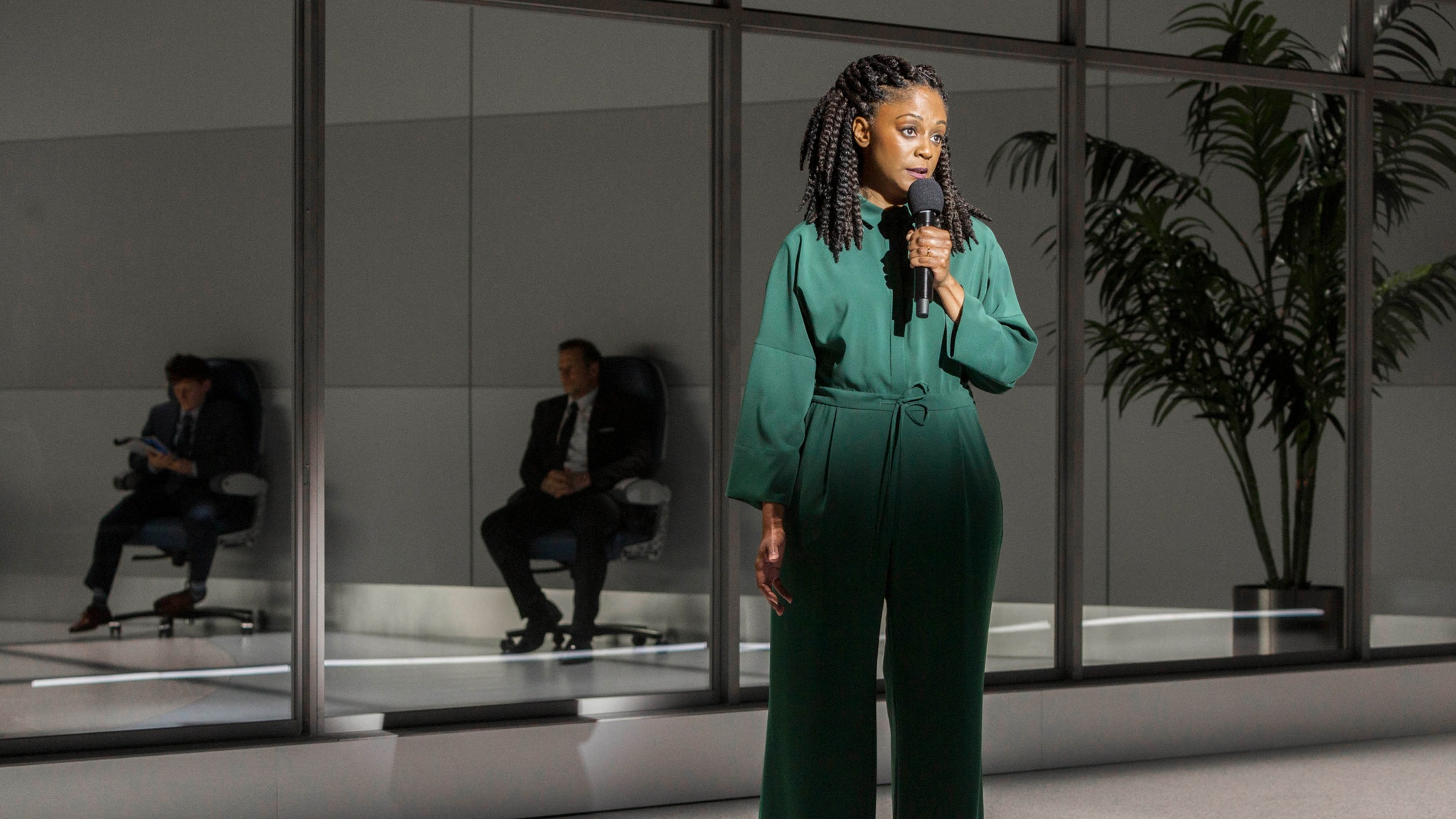 A Black woman in a green jumpsuit stands in front of a stage set resembling an airport waiting room