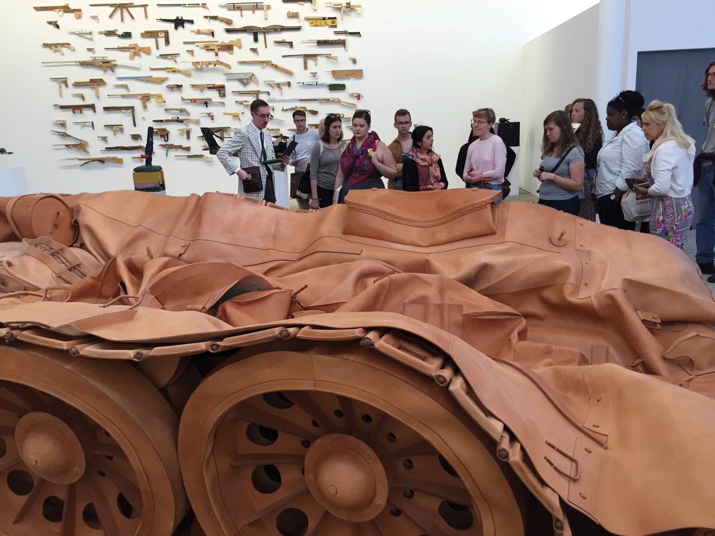 Sculpture of a military tank made of brownish-red leather; the wheels are prominent, but the top of the tank is deflated. Students are standing in the gallery looking at the tank; a multi-piece artwork installation is on the wall behind them.