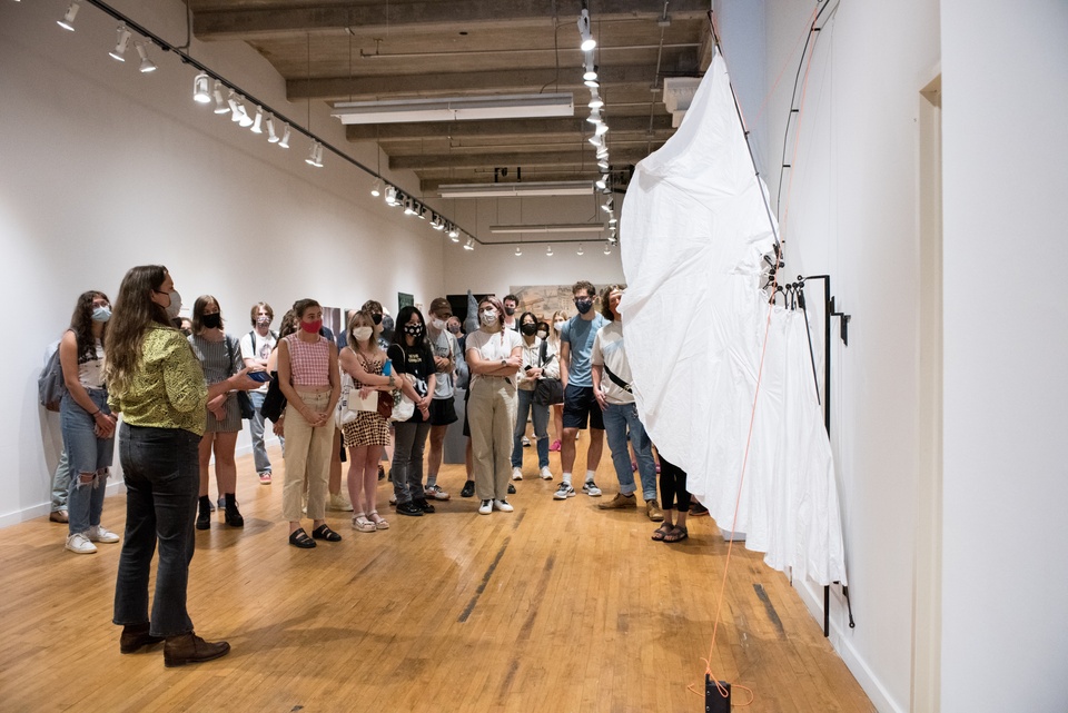 Artist Lindsey stands in front of her large sculpture surrounded by gallery viewers