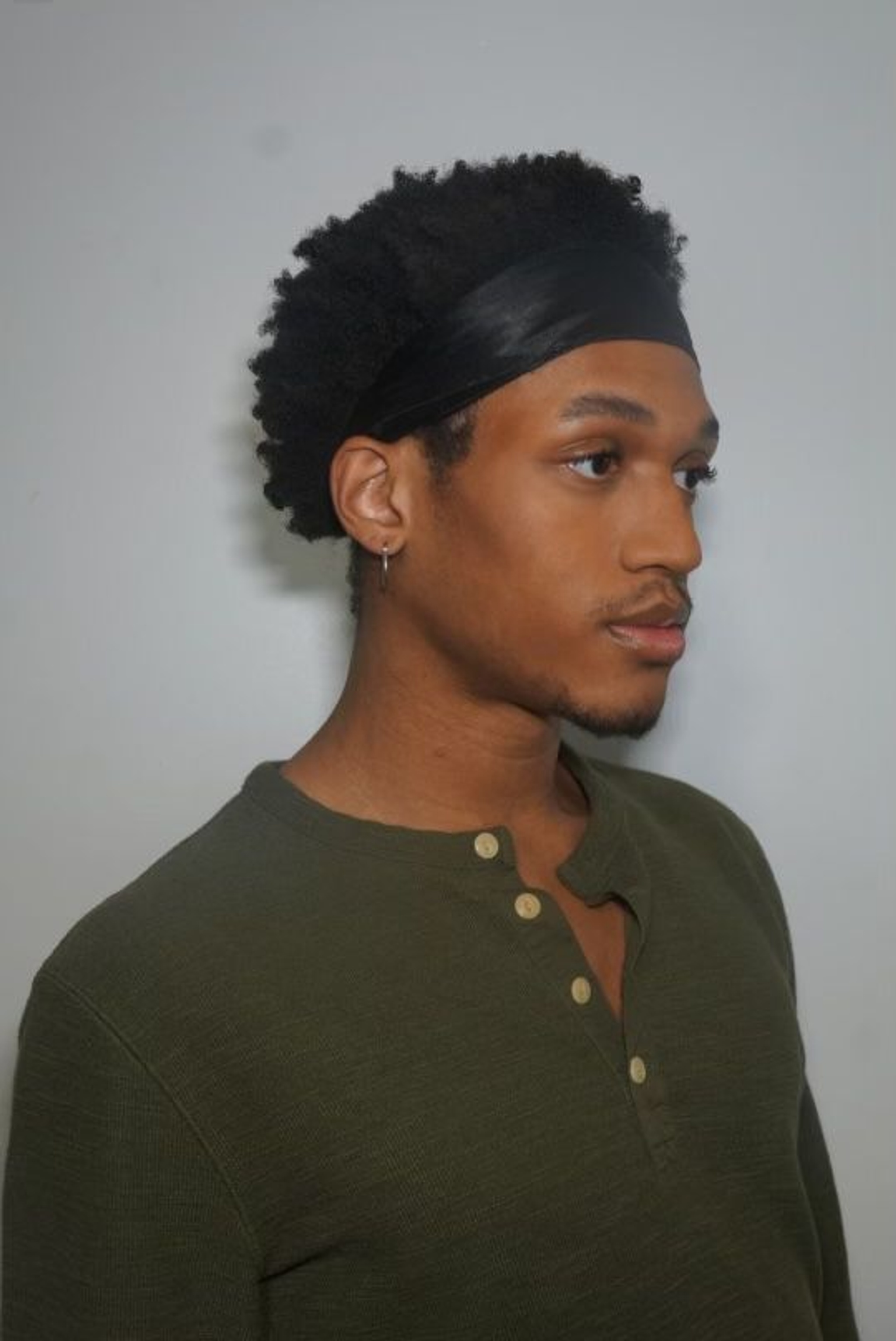 A portrait of dancer Jai Ali McKoy, a Black person with their short hair pushed back with a headband. They look off to the side and wear a dark green henley. 