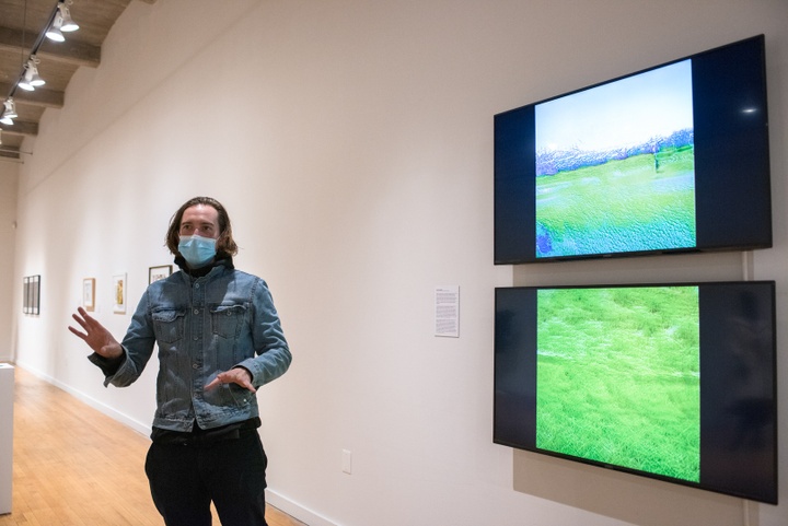Person stands in front of two wall-mounted video screens in a gallery displaying square shaped digital landscape images.
