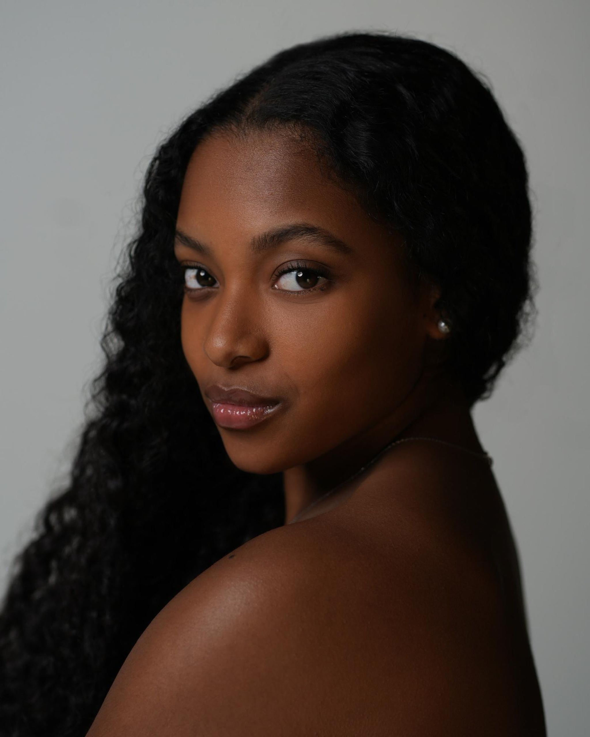 A headshot of dancer Arianna Carson, a Black woman who looks at us from over her bare shoulder. She has long wavy hair pulled around her head to hang down over her right shoulder. 