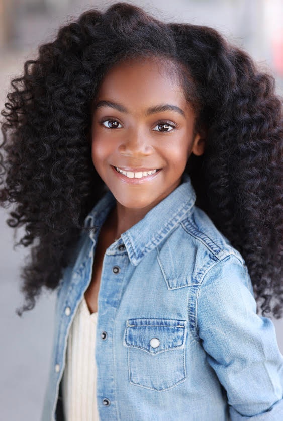 A headshot of child actor Braxton Paul, a young Black girl with curly hair who smiles openly at the camera. She wears a light blue denim jacket. 