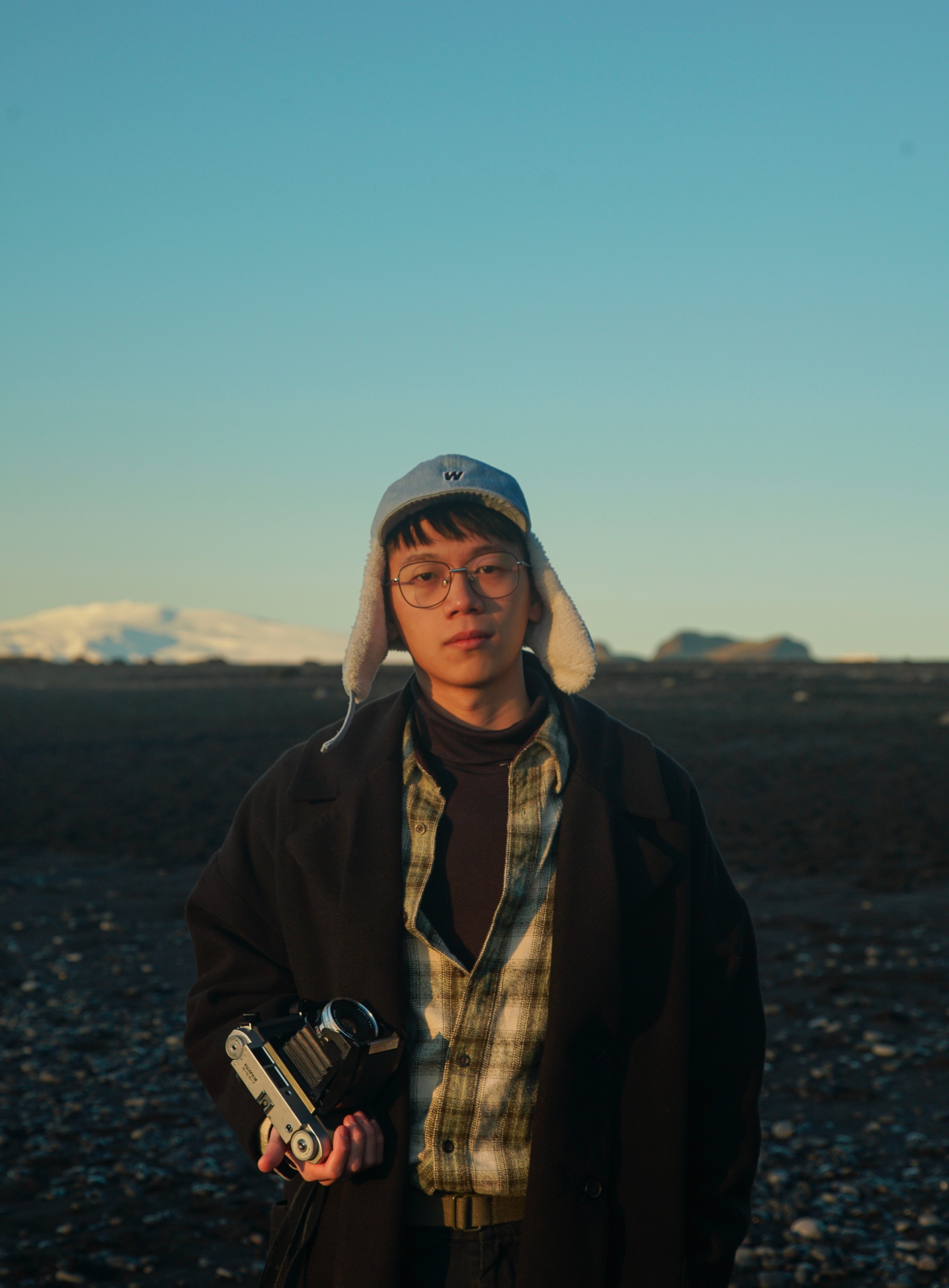A portrait of a young Chinese American man wearing a jacket, plaid shirt, and hat with ear flaps while holding a camera at his hip. He stands against a vast landscape with a white mountain on the horizon in the distance.