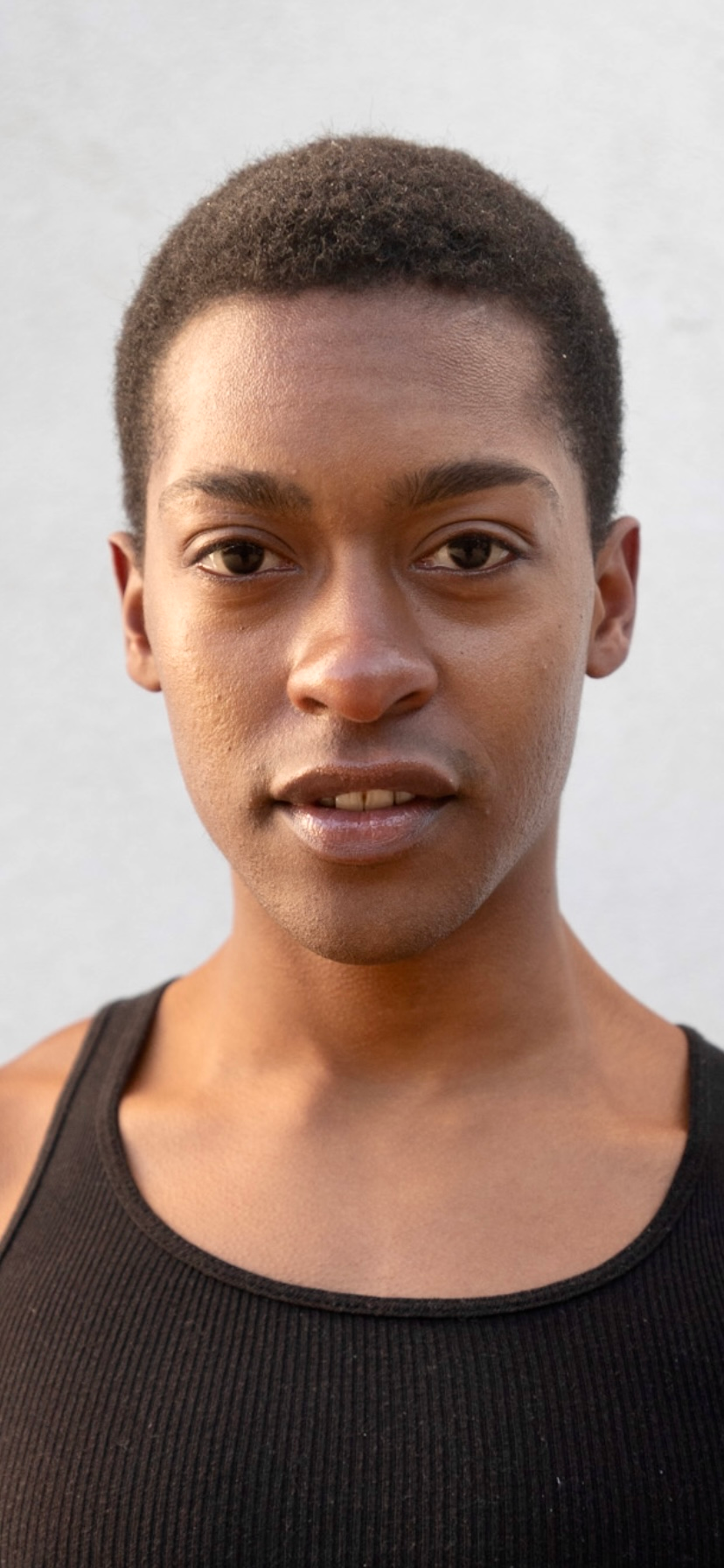 A Black person with short hair wearing a black tank top and standing against a neutral background.