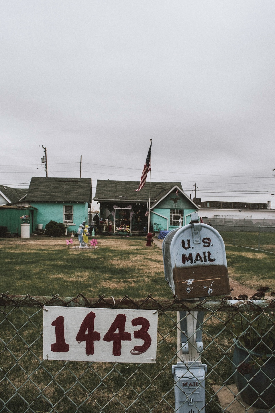 Color photo showing a US Mailbox in front of a small house with bluegreen walls. The number 1443 is on a sign to the left of the mailbox, attached to a fence.