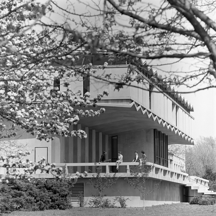 Black and white photo of the corner angle of a midcentury modern building with a large patio. Four people are perched on the patio railing, talking.
