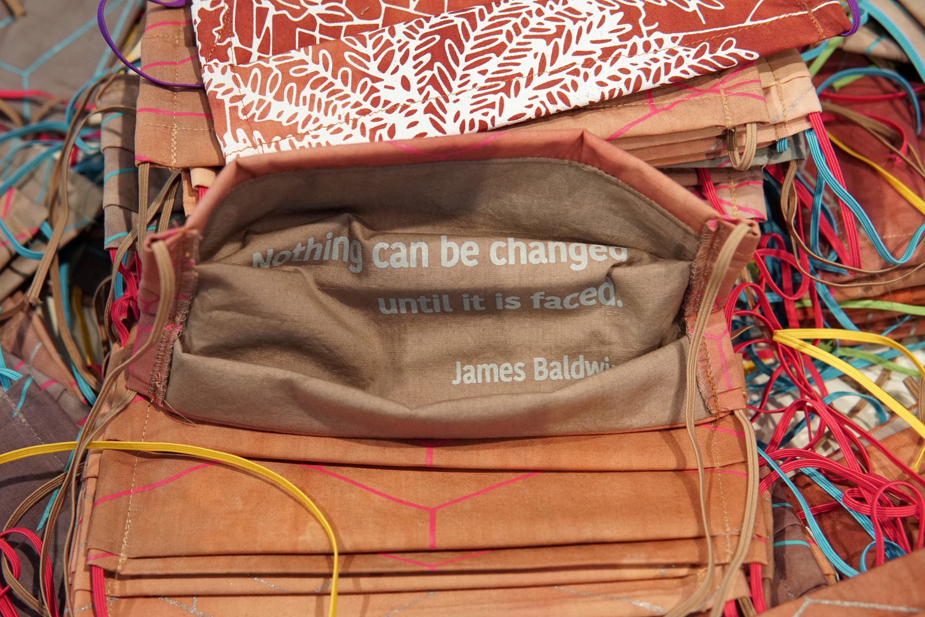 Numerous, colorful, homemade patterned masks laid out. The center mask displays a quote that states, "Nothing can be changed until it is faced. James Baldwin."