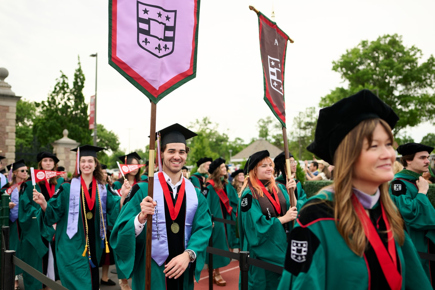 students walking together smiling wearing regalia and holding an Architecture and Art banners at 2026 commencement