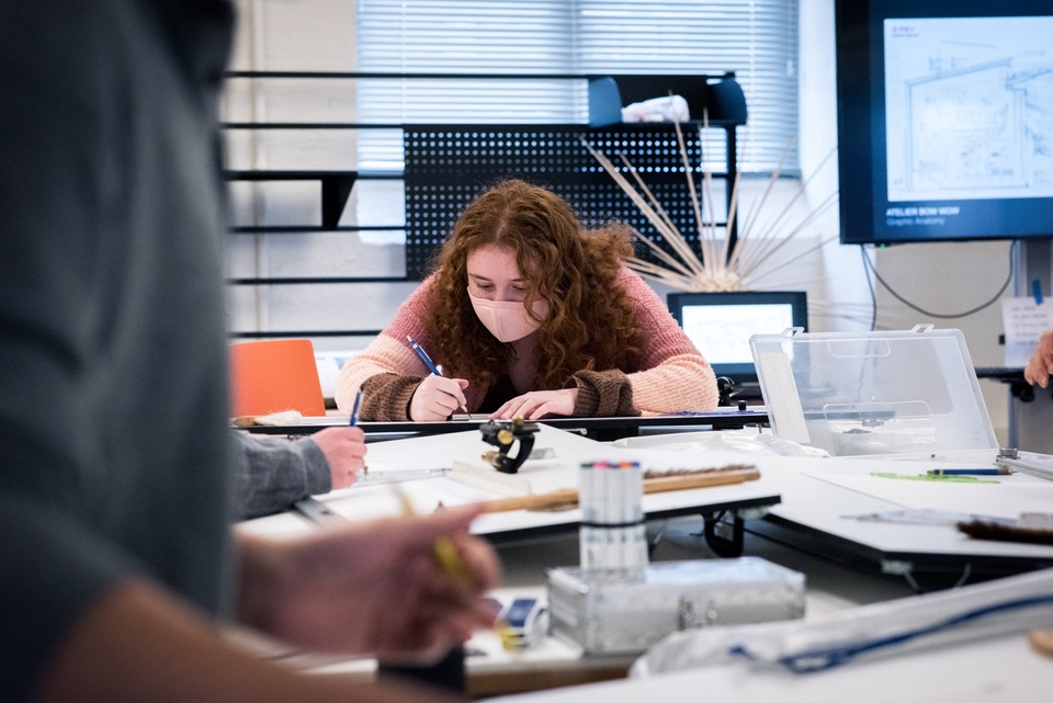 Person bends over a drawing on a drafting table. In the foreground are other drawings, and in the background is a tv screen displaying a complex-looking architectural draft.