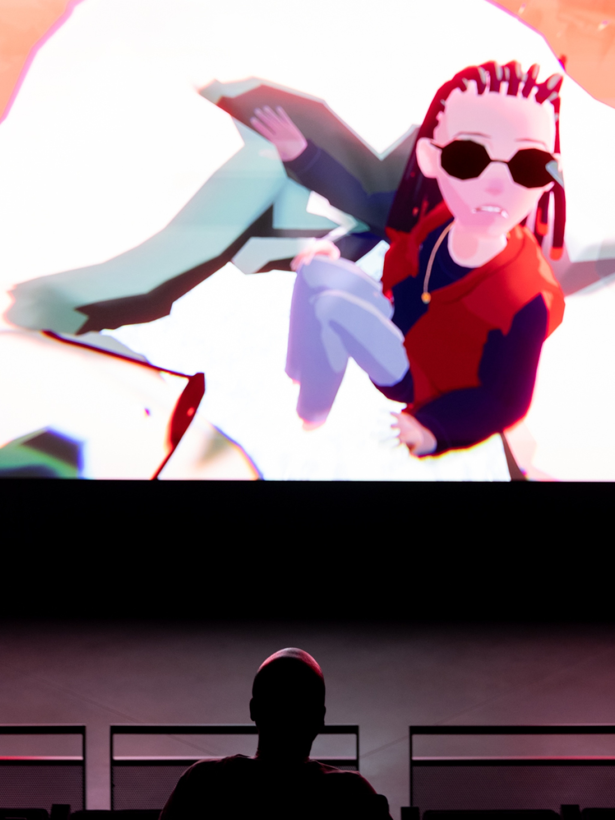 A film screen with a scene from an anime projected on it, with in the foreground the back of a viewer's head pictured from higher up in the cinema-style seating