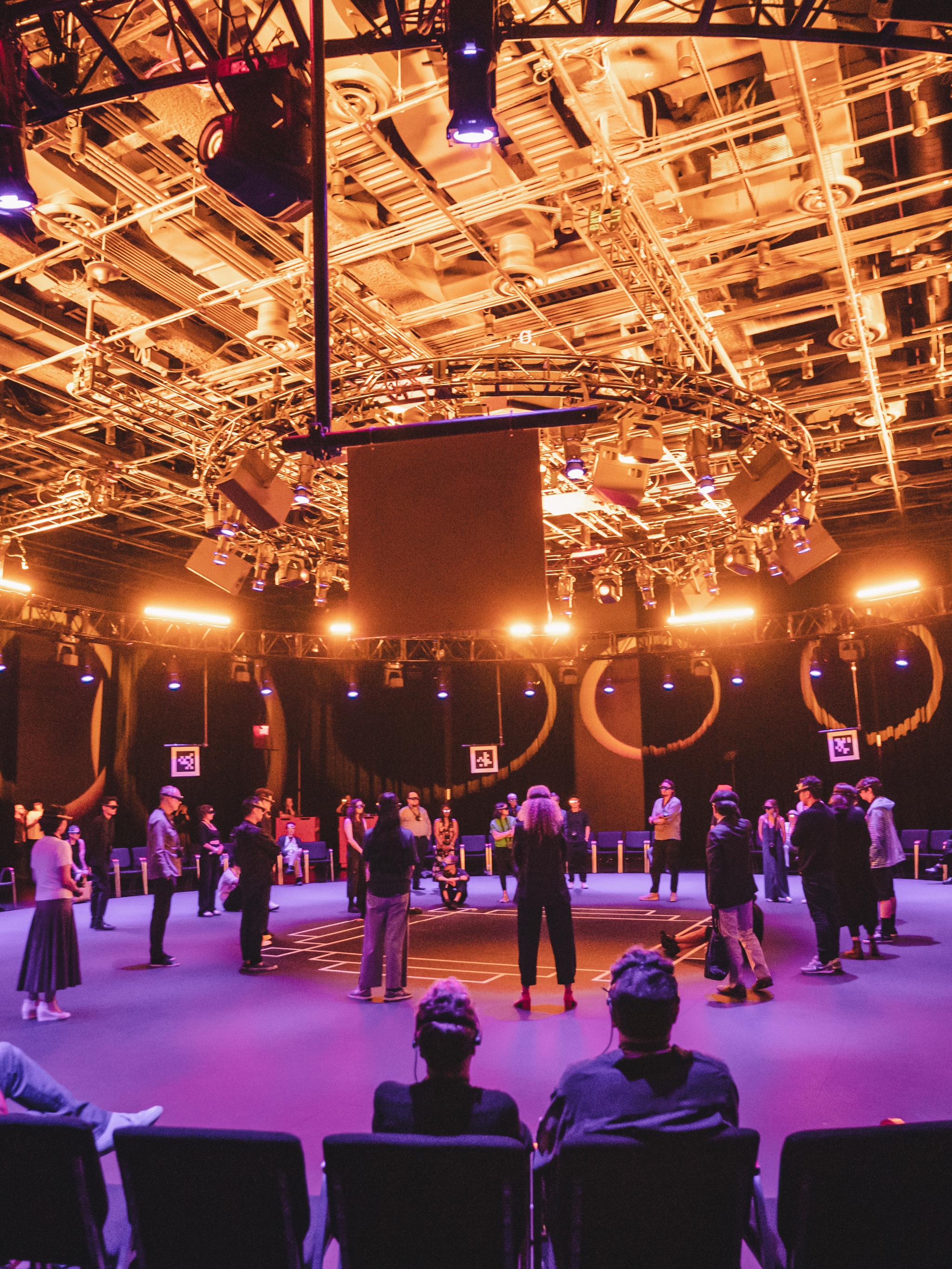 In a theater space arranged with a wide circle of chairs facing inside the circles, audiences gather in the center of the space, standing and crouching. They were headsets that look like googles over their eyes. The space is lit in purple light that illuminates the floor and a bright golden light that reveals the infrastructure of pipes and ducts in the ceiling.