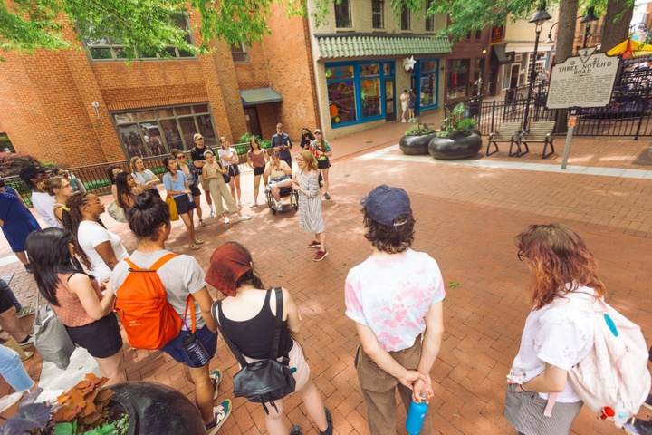 People gather around an individual speaking animatedly within a brick-lined plaza with shops in the background. The historic sign in the background reads Three Notch D Road.