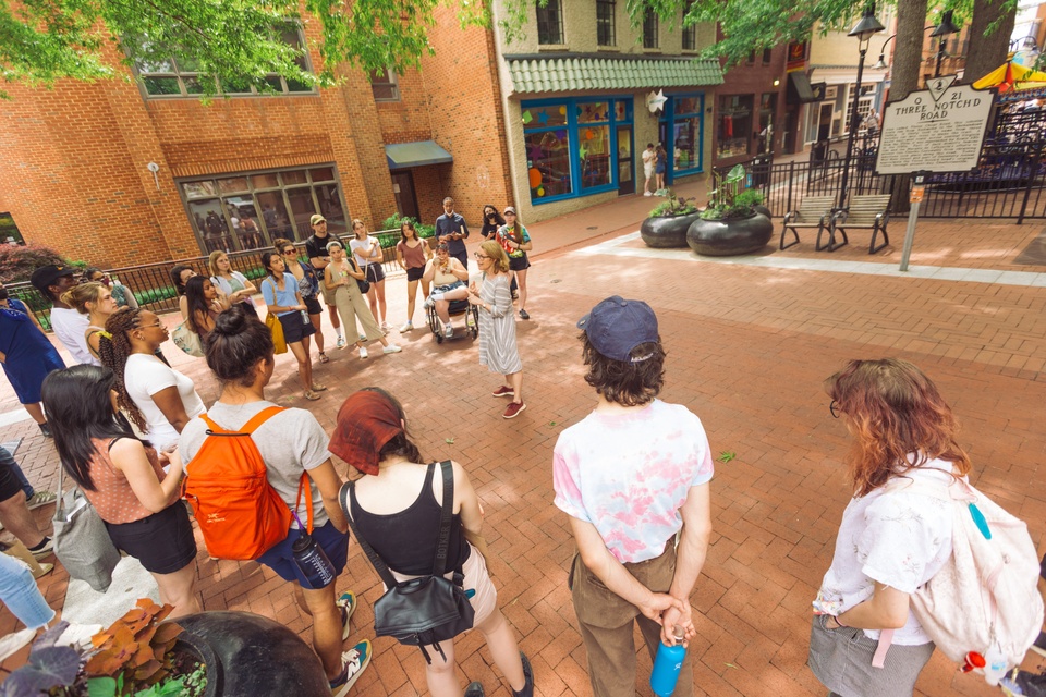 People gather around an individual speaking animatedly within a brick-lined plaza with shops in the background. The historic sign in the background reads Three Notch D Road.