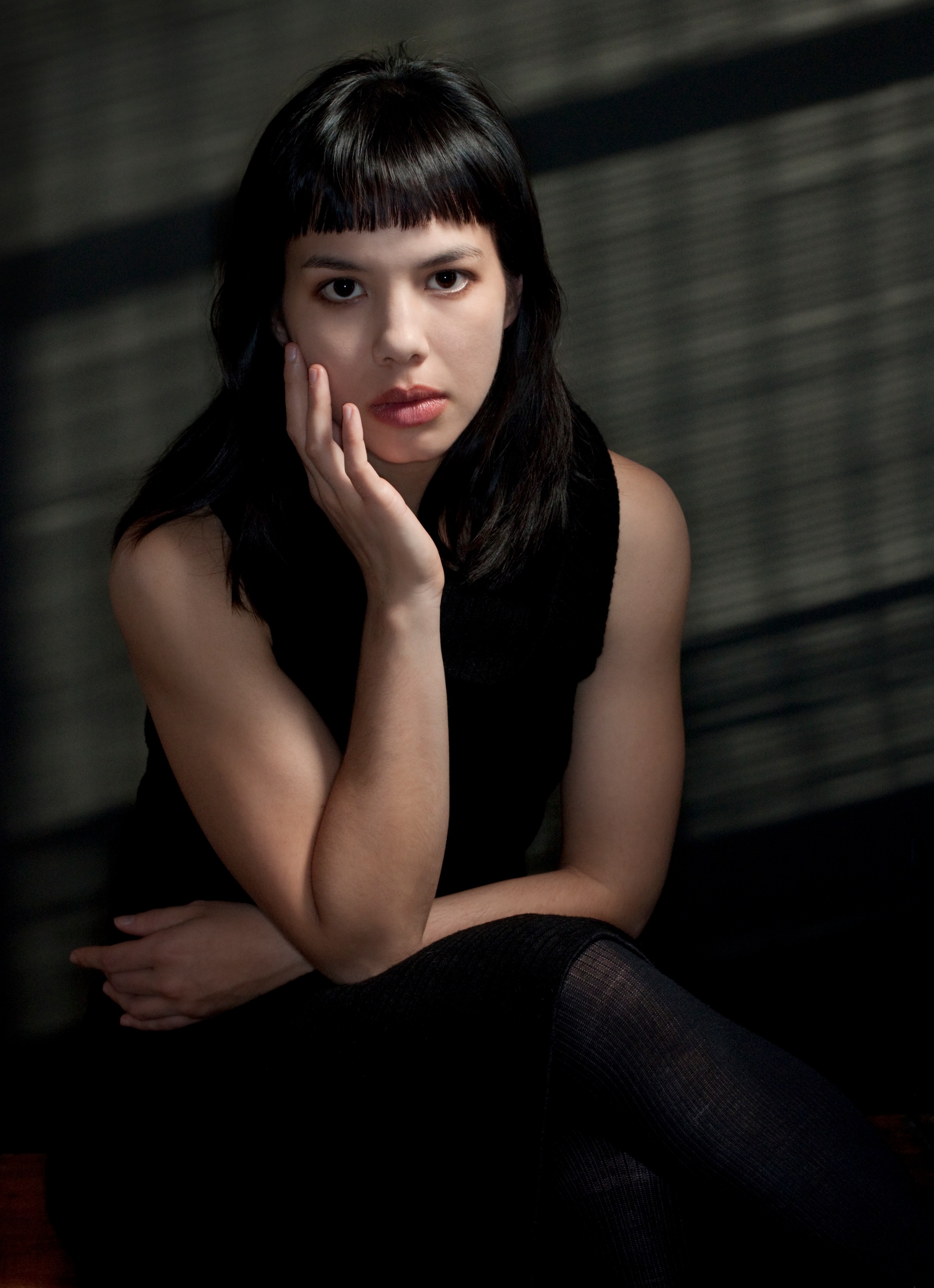 A portrait of dancer Pareena Lim sitting with legs crossed, one arm lifted so that her right cheek rests on her hand. 