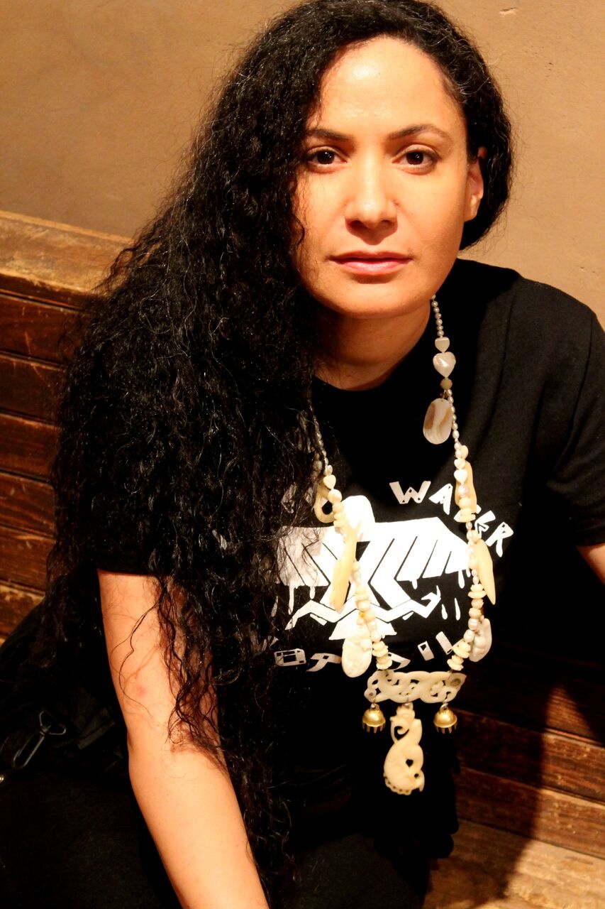Singer and composer Jennifer Kreisberg sits on a bench. She has long, wavy dark hair and wears a long necklace over a black t-shirt. 