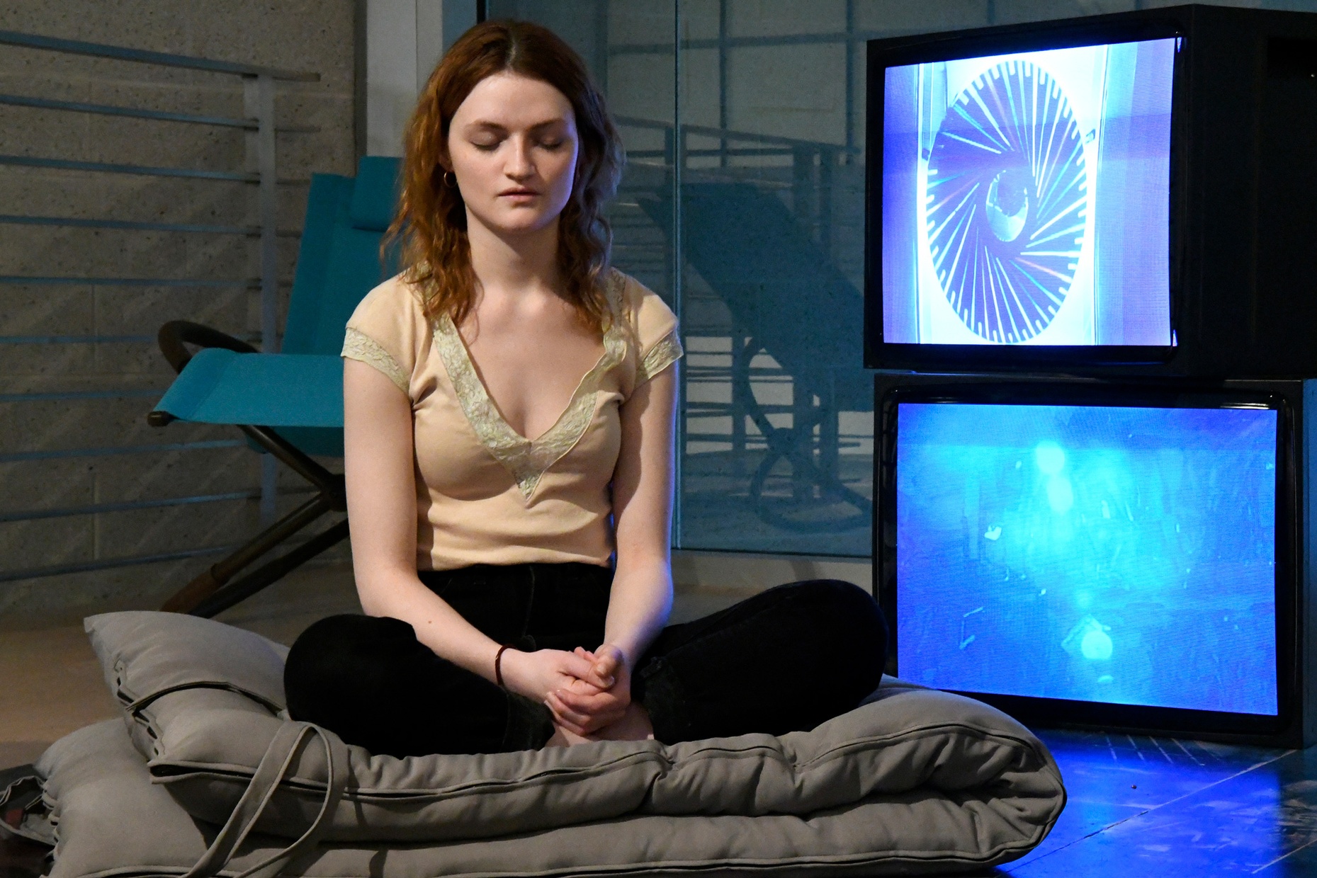 A light-skinned, young female sits with eyes closed and cross-legged on a square gray pillow in front of two TV's stacked on each other.