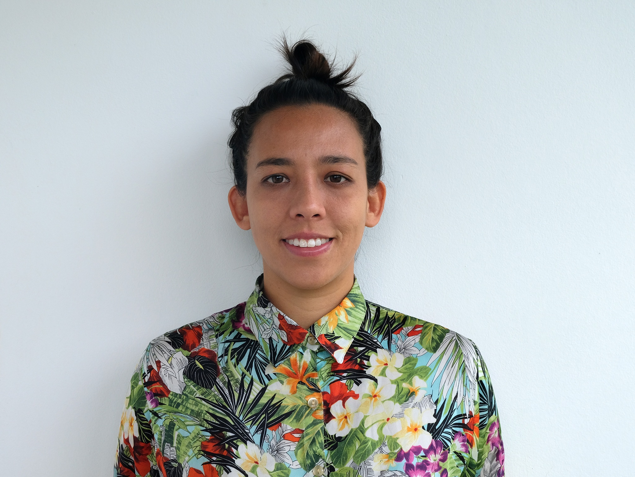 A portrait of curator Valentina Tong shown from the shoulders up against a blank, white wall. Valentina has dark hair pulled back into a loose bun at the top of her head. She smiles, looking straight ahead, and wears a colorful floral-print button down shirt buttoned to the collar.