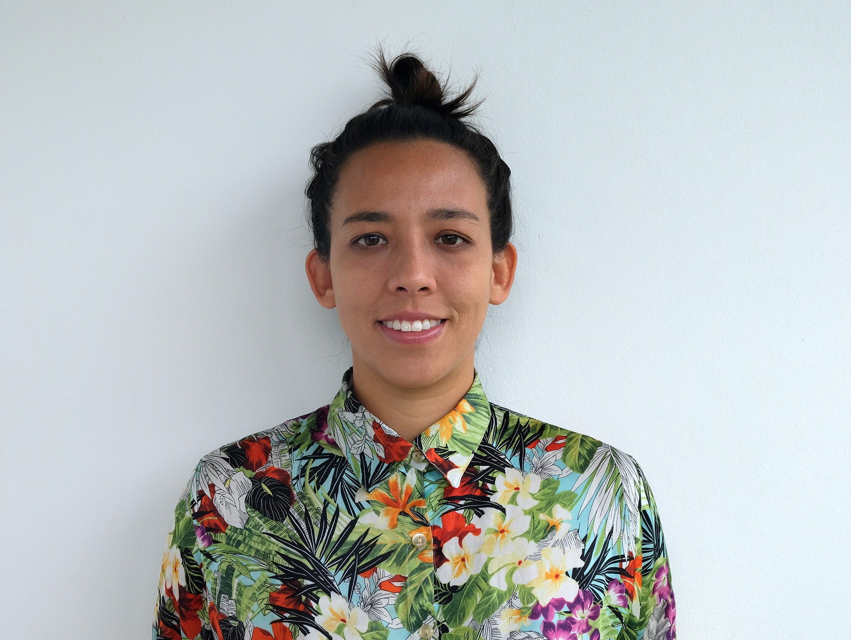 A portrait of curator Valentina Tong shown from the shoulders up against a blank, white wall. Valentina has dark hair pulled back into a loose bun at the top of her head. She smiles, looking straight ahead, and wears a colorful floral-print button down shirt buttoned to the collar.