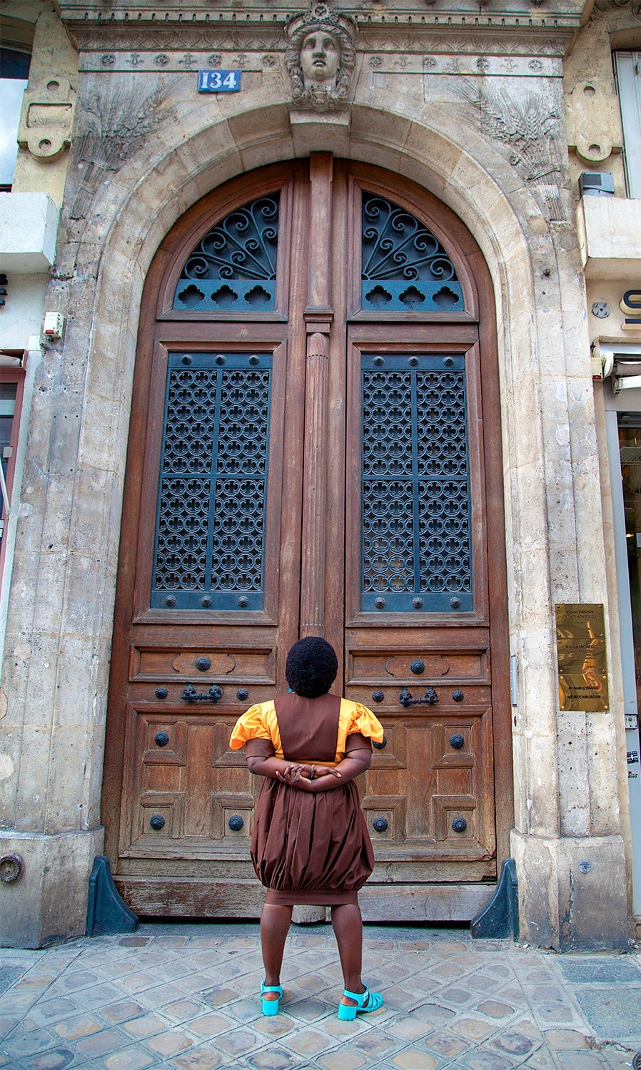 Photo of an individual with their back to the camera, stairing at a massive, curved wooden double door with glass inlay. Stonework frames the door.