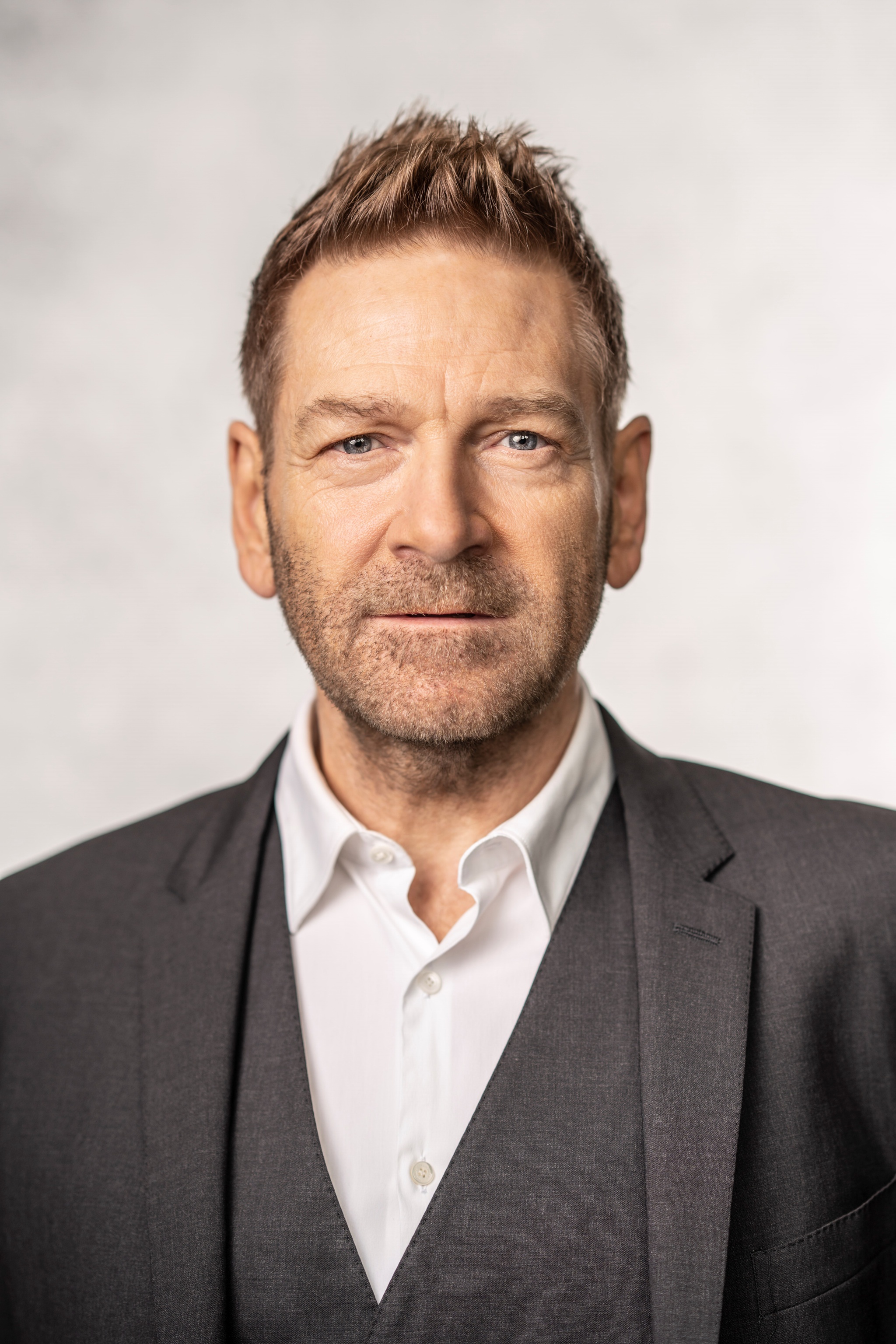 A headshot of actor Kenneth Branagh, a white man with light brown hair. Kenneth looks directly at us and wears a three piece suit, without a tie. 