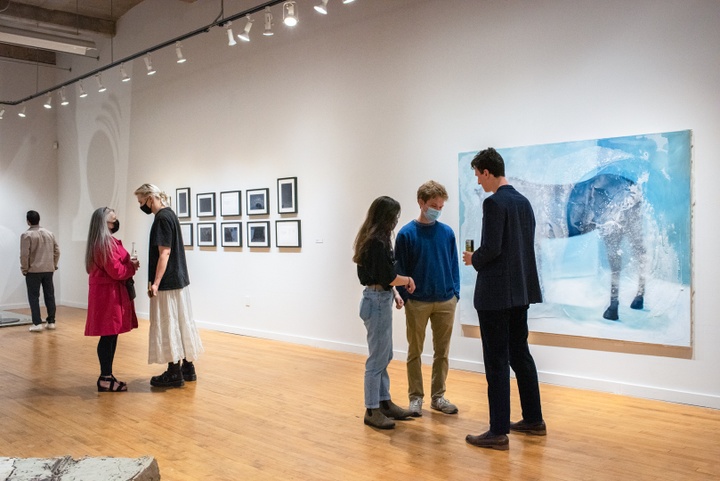 Several groups of people stand in a gallery space showcasing a blue and grey abstract painting of a horse and a series of small framed pieces in black and white.
