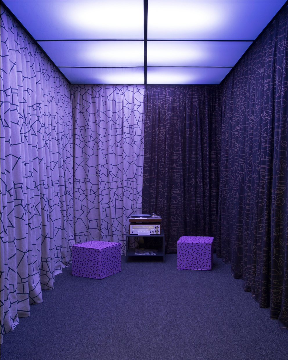 the Tang elevator enclosed with floor-length curtains hanging on all of the walls. Two cube seats are positioned next to a record player and receiver on a stand. The lights in the ceiling are purple, casting purple light on everything