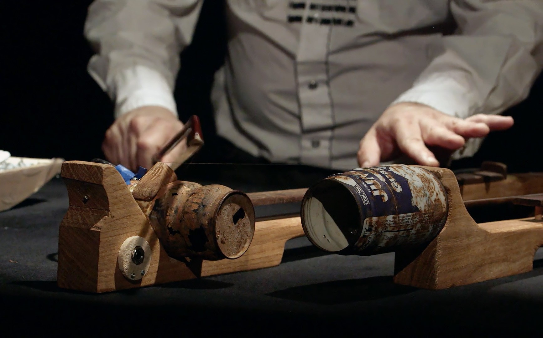 A handmade string instrument made of wood and old cans sitting on a table being played by a light-skinned person wearing a white, button-up shirt holding a violin bow.
