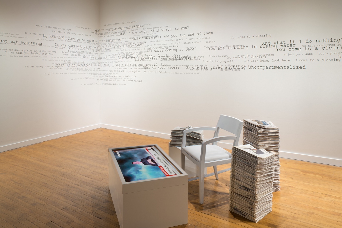 Installation view of a gallery space: in the foreground, a chair next to a video on a screen in a white platform/box, with stacks of newspapers surrounding this all. In the background, wires from exposed machinery and other artworks mounted to the wall. The walls are white with some text running along it in a typewriter font. The floors are wooden.